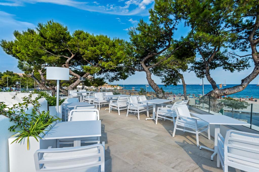 Terases zona ar baltām mēbelēm, kas atrodas pie jūras, apstādīta ar zaļiem kokiem un skatu uz pludmali.