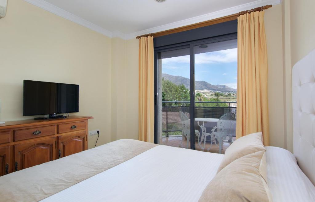 Mūsdienīga guļamistaba ar skatu uz kalniem, kurā ir televizors, koka mēbeles un balkons ar baltiem dārza krēsliem. Gaismas krāsas interjers rada mierīgu atmosfēru.