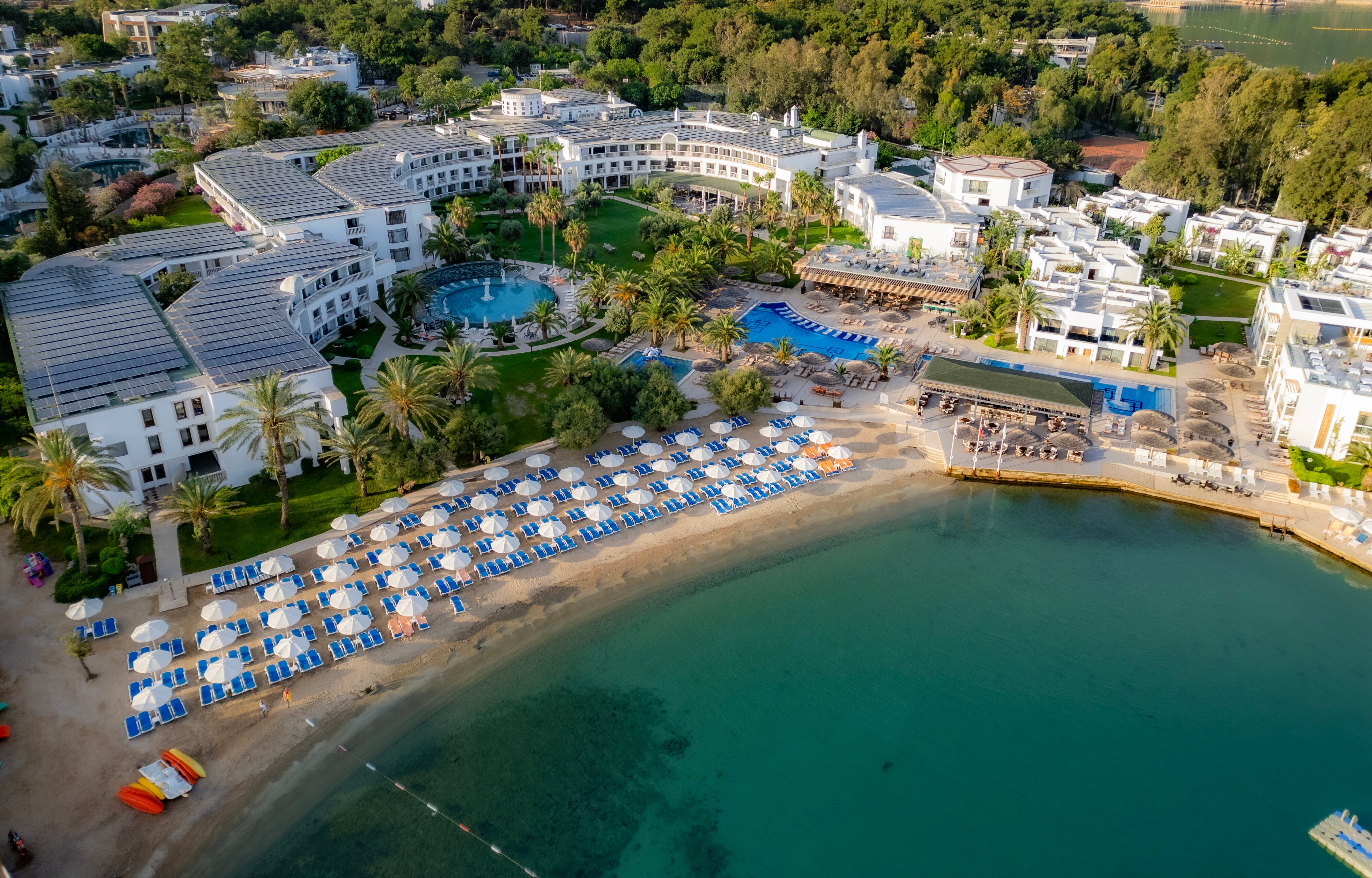 Viesnīcas komplekss ar baseiniem un pludmali, kurā ir saulessargi un sauļošanās krēsli. Apkārt ir zaļš parks ar palmām un ainavisku ūdenstilpni. panoramic view with beach and pools