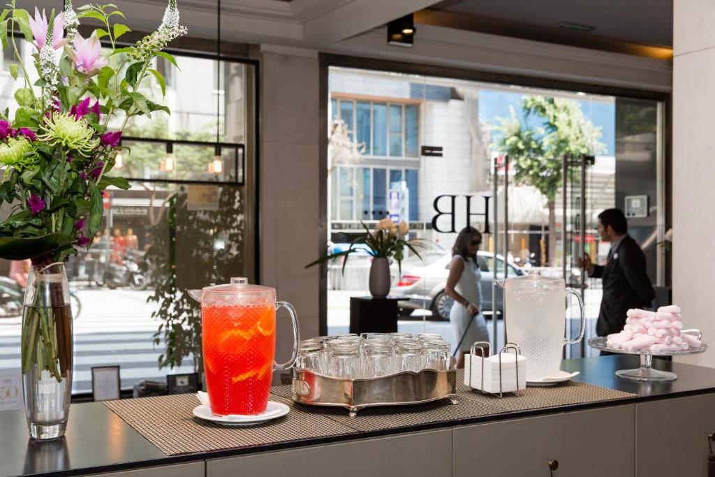 Viesnīcas reģistratūra ar ziedu kompozīciju, divām dzērienu krūzēm un saldumiem uz galda, caur logu redzama pilsētas aina.