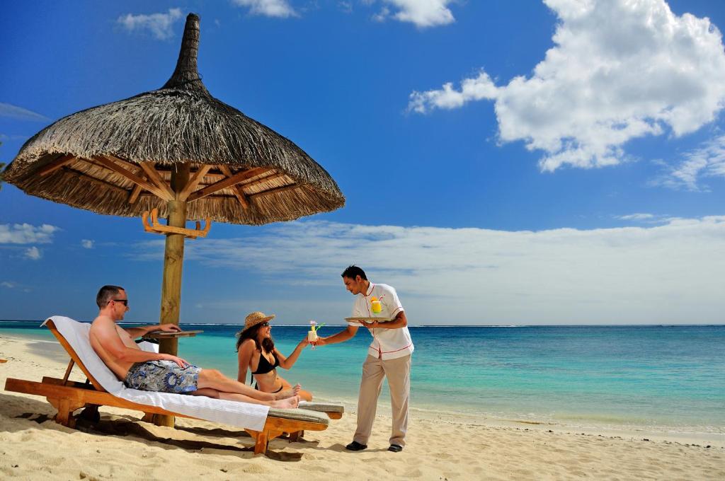 Pludmales ainava ar cilvēkiem, kas atpūšas zem salmu jumta un saņem dzērienus no apkalpojošā personāla. Skats uz skaisto jūru un skaidro debesi.