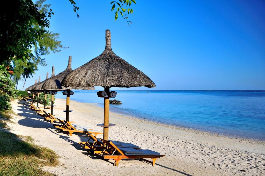 Pludmales ainava ar saules aizsargiem un koka atpūtas krēsliem, kas izvietoti gar smilšu krastu pie skaidra, zila ūdens.