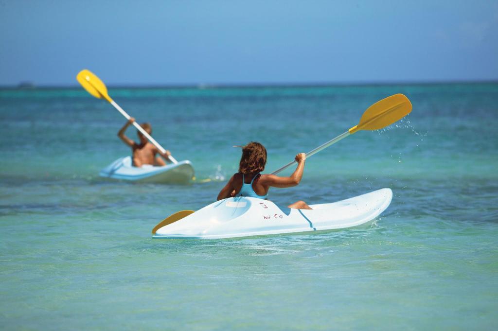 Divi cilvēki, kas airē kajakā, baudot saulainu dienu pie krasta ar dzidru, zilu ūdeni.