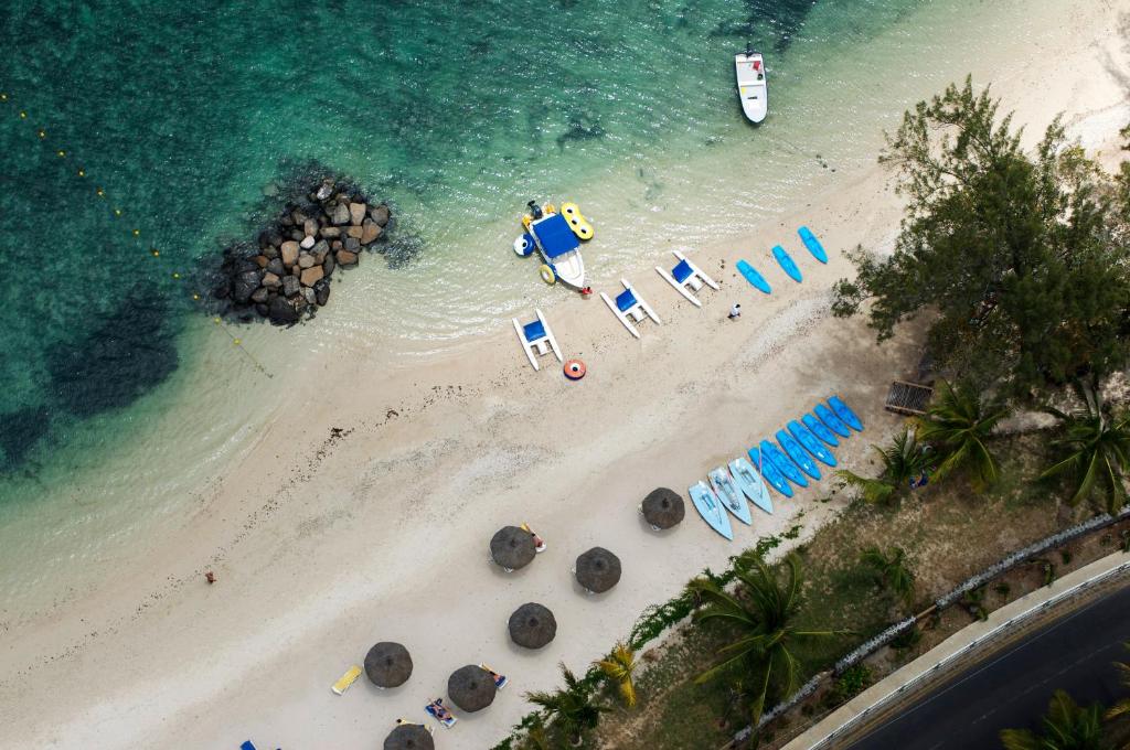 Pludmales skats no augšas ar baltu smilšu krastu, zilām laivām un saulessargiem. Skaidra zila ūdens virsma un zaļie koki papildina relaksējošo atmosfēru.