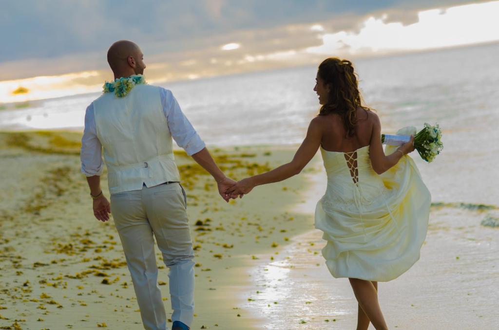 Jaunais pāris, tērpies kāzās, pastaigā pa pludmali, turot viens otra roku. Saule noriet, radot romantisku atmosfēru.