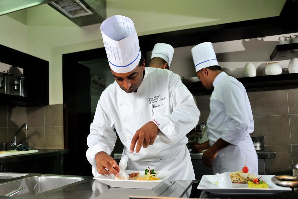 Šefpavārs rūpīgi izdekorē ēdienu restorāna virtuvē, kamēr citi pavāri strādā fonā. Šī aina atspoguļo profesionālo kulināriju un radošo pieeju ēdienu pagatavošanā.