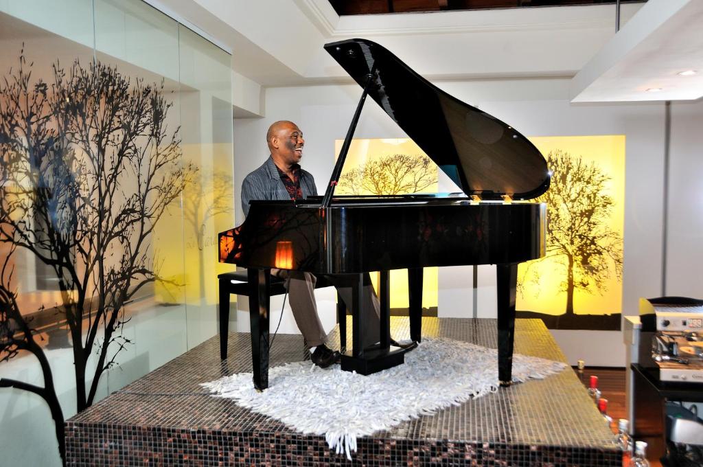 Vīrietis spēlē melno klavieri stilīgā interjera telpā, kurā redzamas stilizētas koku ilustrācijas uz sienām. Gaisma rada siltu atmosfēru, pievienojot mājīgumu.