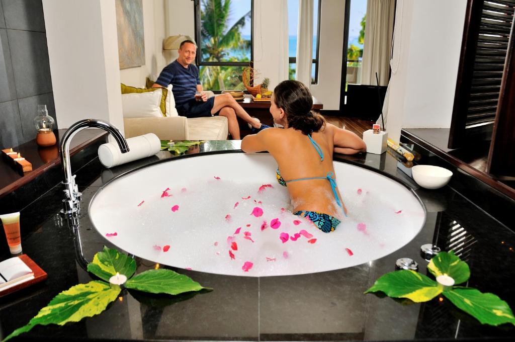 Relaksējoša ainava luksusa viesnīcas numurā ar burbuļvannu, kurā ir ziedlapiņas, un personu, kas atpūšas blakus. Ievērojama tropiskā atmosfēra un miera sajūta.