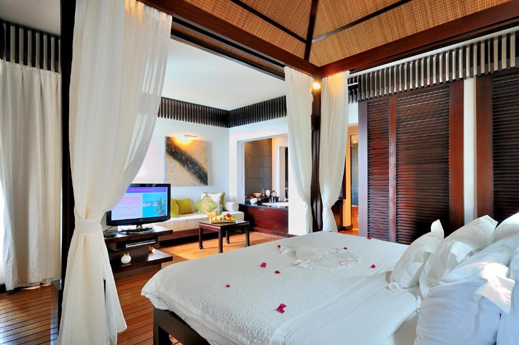 Eleganta viesnīcas numura interjers ar gultu, rotātu ar ziedlapiņām, un komfortablu atpūtas zonu. Istabā ir mūsdienīgs televīzijas ekrāns un mājīga atmosfēra.