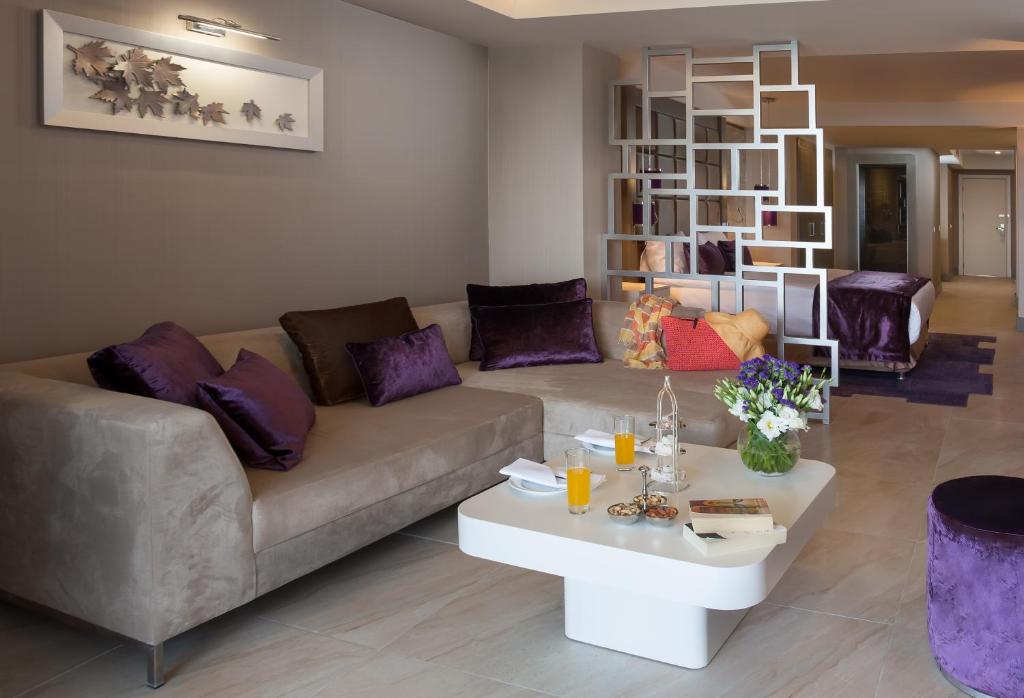 Mūsdienīgs viesnīcas numurs ar elegantu viesistabu, kurā ir pelēka dīvāna un violeti spilveni, kā arī stilizēts atdalītājs un galda dekorācijas. Interjers ir gaišs un gaumīgs, radot patīkamu atmosfēru.