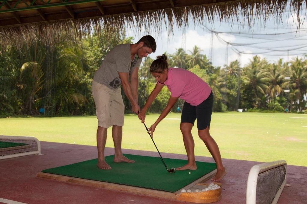 Divi cilvēki nodarbojas ar golfa apmācību, viens palīdzot otram uzlabot sitiena tehniku. Fons ir zaļš golfa laukums ar palmām.