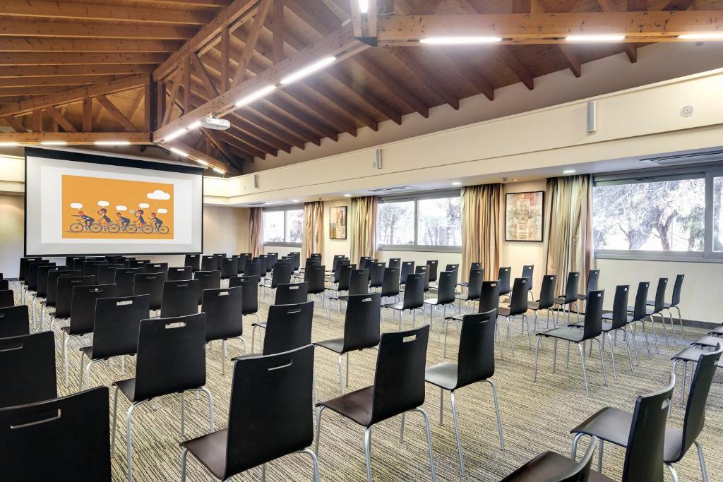 Konferenču zāle ar melniem krēsliem un projektoru, kas rāda ilustrāciju ar cilvēkiem, kas brauc ar velosipēdiem. Telpa ir gaiša un plaša, ar koka griestiem un logiem, kas nodrošina dabisko apgaismojumu.