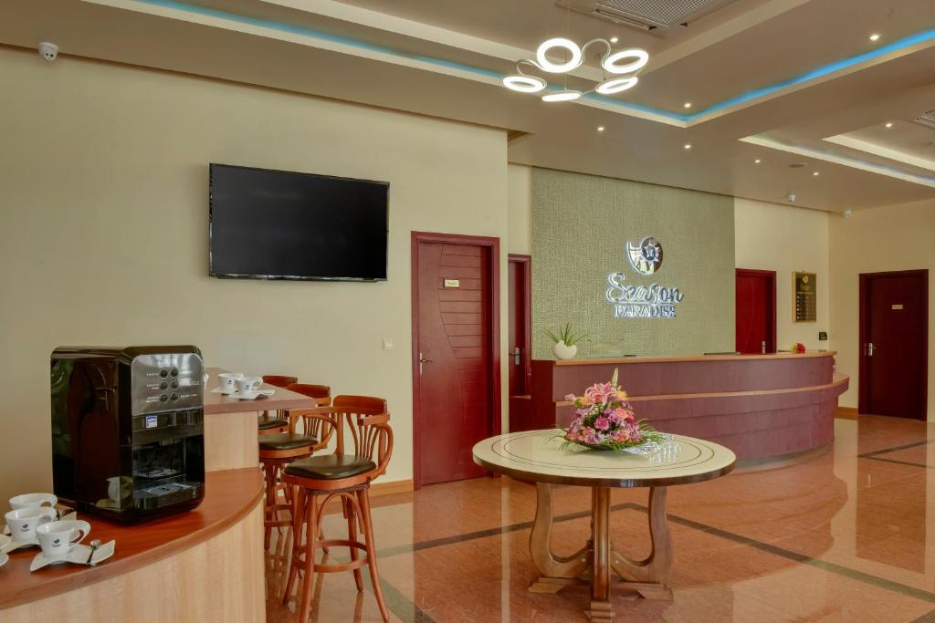 Mūsdienīgs viesnīcas recepcijas interjers ar koka mēbelēm, kafijas automātu un ziedu kompozīciju uz galda. Uz sienas redzams televizors un viesnīcas logo.