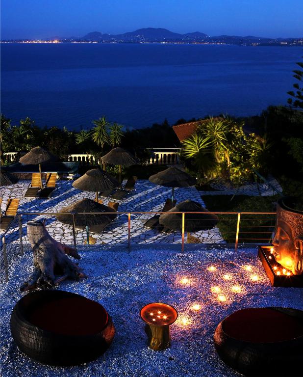 Elegantā nakts ainava pie jūras, kur redzami atpūtas krēsli, palmām un lampiņām apgaismota terase. Skats uz mierīgu ūdeni un kalniem fona.