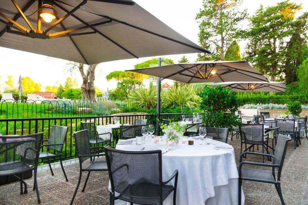 Āra restorāna terase ar galds, ērtām krēslām un saulessargiem, apzaļumojums fonā. Ideāla vide relaksācijai un ēdiena baudīšanai.