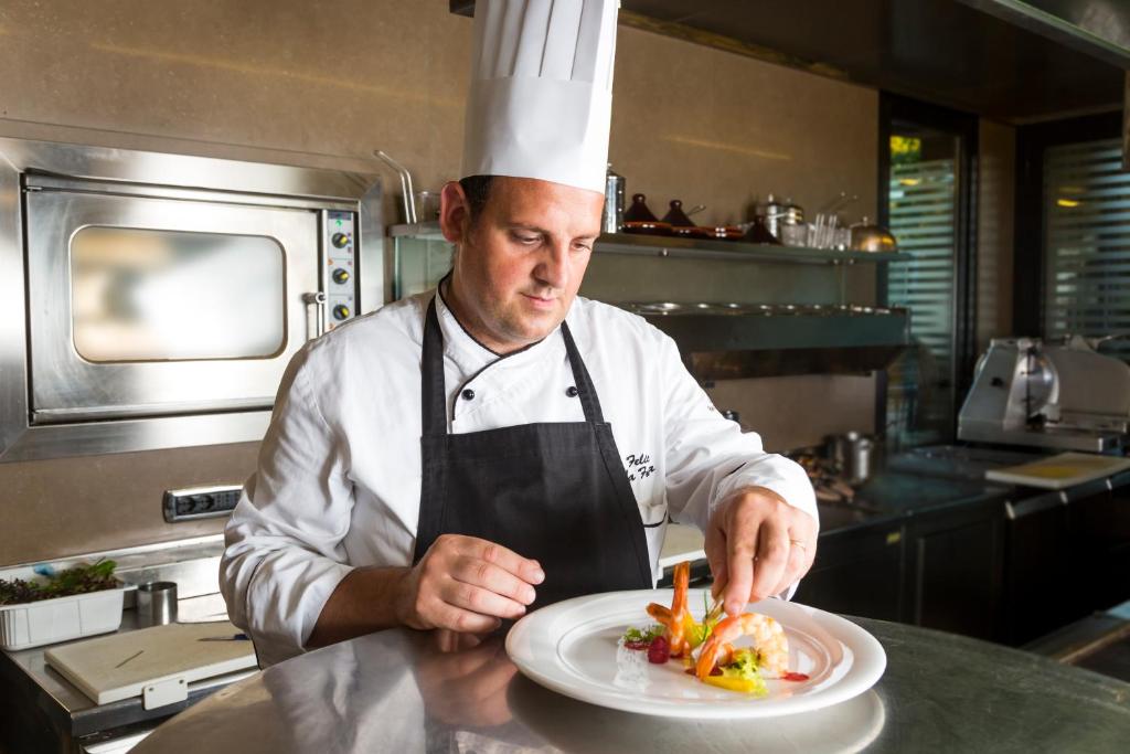 Šefpavārs ar balto cepuri rūpīgi dekorē plāksni ar ēdienu, izmantojot svaigas sastāvdaļas. Fons parāda mūsdienīgu virtuvi ar gatavošanas aprīkojumu.