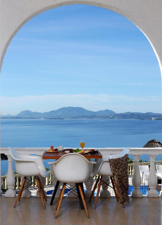 Skats uz jūru no terases ar galdu, kurā ir ēdieni un dzērieni, un ap ēdamgaldu ir modernas krēsli. Fons ir kalnu siluets un skaidrs debesis.