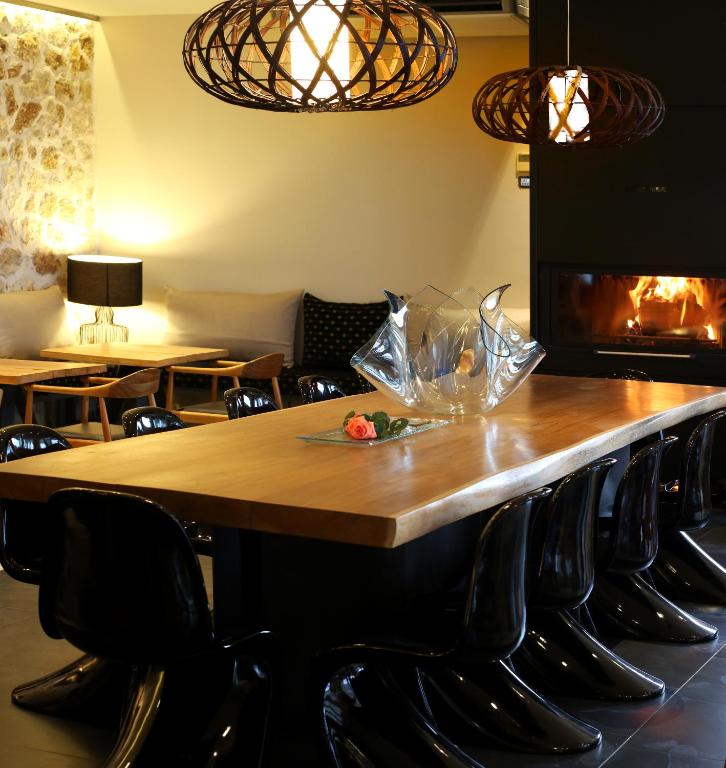Mūsdienīgs interjers ar lielu koka galdu un melnām krēsliem, apgaismots ar stilīgām lampām. Uz galda ir dekoratīvs trauks ar ziedu, blakus redzama kamīna liesma.