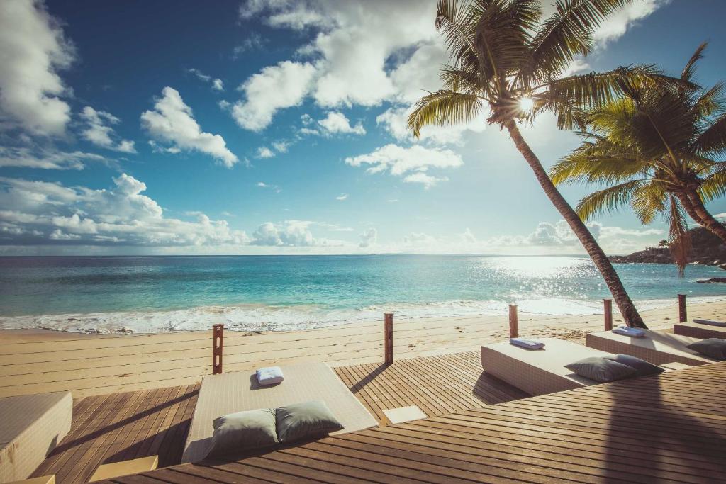 Tropu pludmale ar palmām un saulainu debesu fonu, kurā redzami atpūtas krēsli un mierīga jūra. Ideāla vieta relaksācijai un atpūtai.