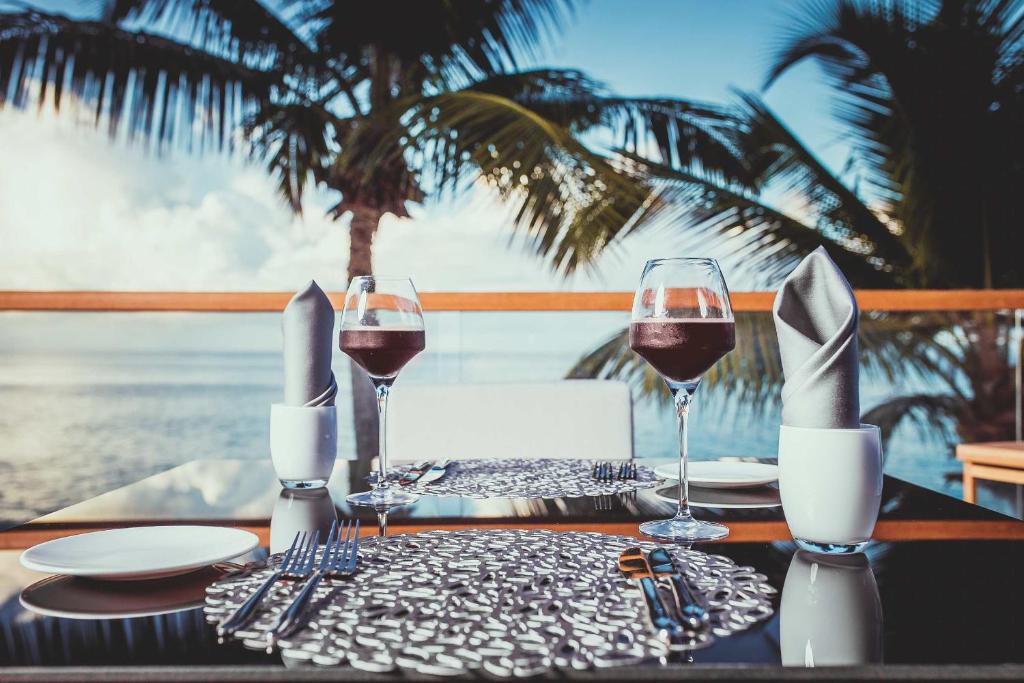 Elegants restorāna galdiņš ar vīna glāzēm, salvešu un trauku komplektu, fona attēlā redzami palmas un jūra. Ideāla atmosfēra romantiskām vakariņām.