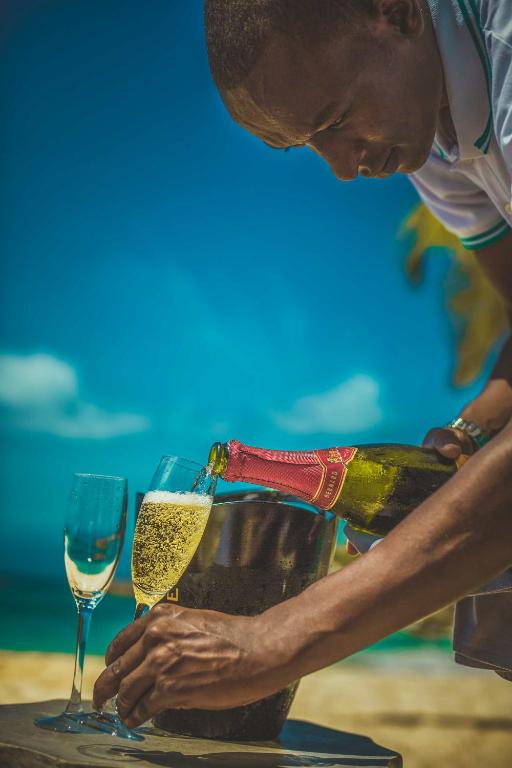 Vīna pudele tiek izlieta divos šampanieša glāzēs, kamēr cilvēks stāv pie pludmales ar skaistu zilo debesu fonu.