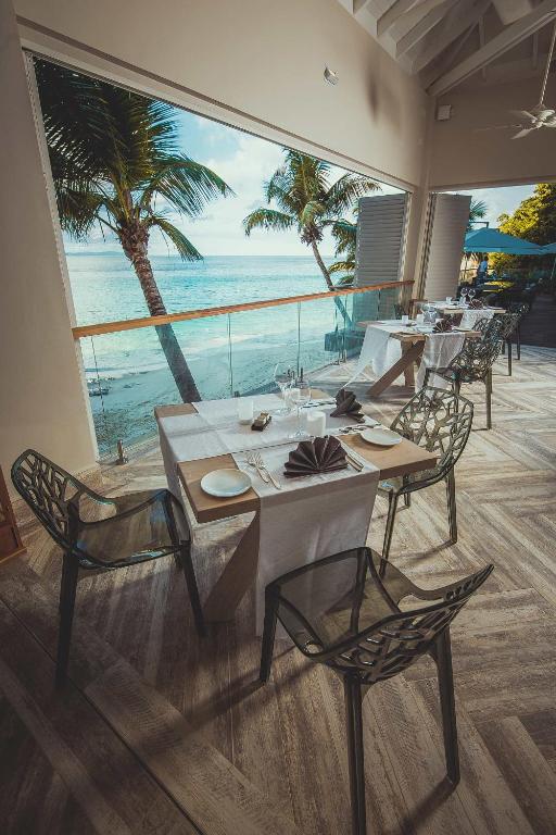 Elegants restorāns ar skatu uz pludmali, rotāts ar mūsdienīga dizaina galdiem un krēsliem, kas aicina baudīt maltīti. Palmām fona, ēdināšanas atmosfēra ir relaksējoša un pievilcīga.
