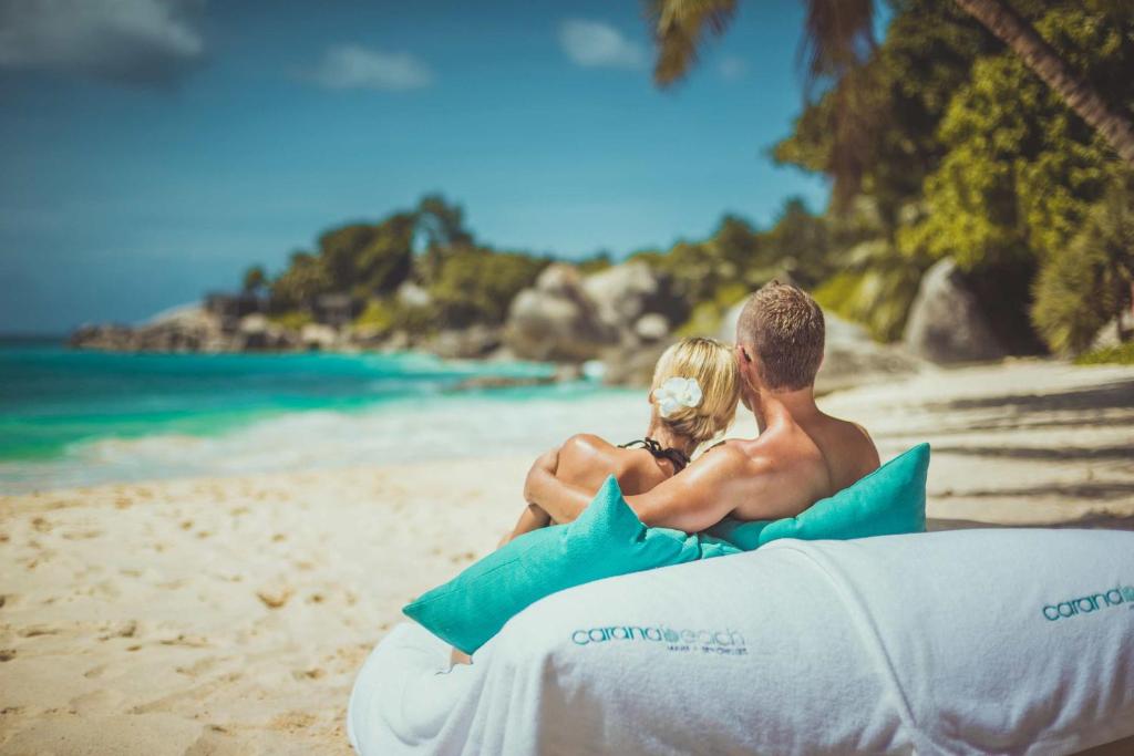 Pāris relaksējas pludmalē, sēžot uz mīksta dīvāna, un skatās uz zilo jūru un saulaino debesīm. Ideāla romantiska brīvdienu aina.