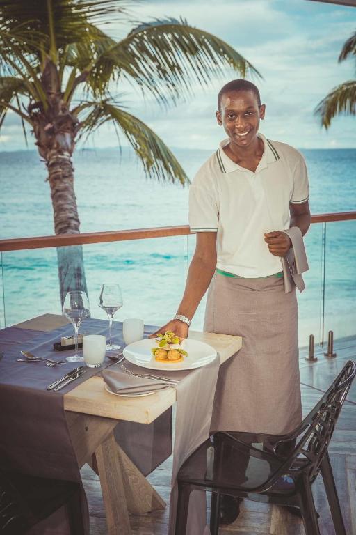 Viesmīlis pasniedz ēdienu pie eleganta galda ar skatu uz jūru, iestatīts pludmales restorānā, kur ir palmas un gleznains fons.