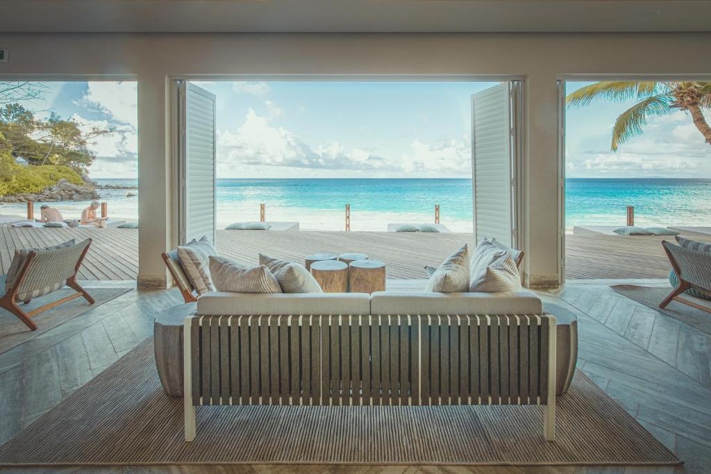 Mūsdienīga viesnīcas telpa ar skatu uz pludmali, kurā ir ērta dīvāna un koka galdiņu. Priekšplānā redzama atvērta terase, kas ved uz zilo jūru.