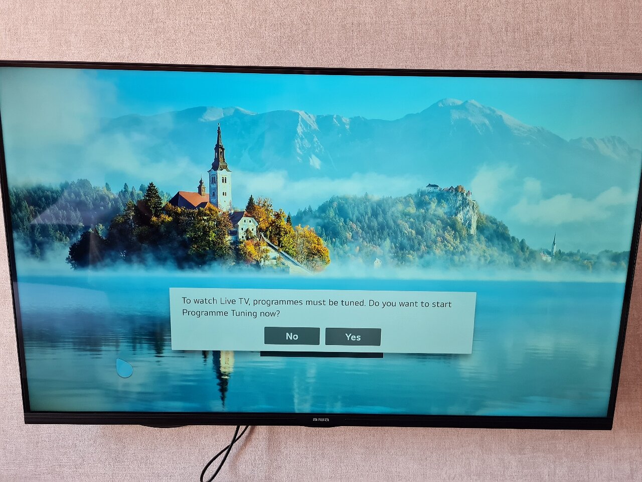Ekrānā redzama ainava ar kalniem un ezeru, kurā atspoguļojas baznīca un meži. Paziņojums informē par nepieciešamību noregulēt programmas, lai skatītu tiešraides televīziju. panoramic view with beach and pools