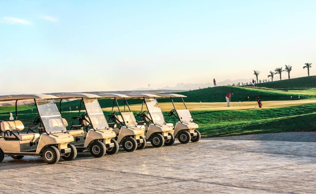 Golfu ratiņi stāv rindā uz gleznainā golfa laukuma fona, kur spēlētāji izpilda sitienus saulainā dienā.