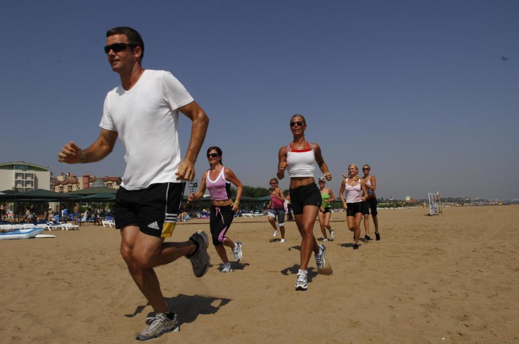 Grupas cilvēku, kas skrien pa pludmali saulainā dienā, ar viesnīcas ēku fonā. Sievietes un vīrietis ģērbti sportiskos tērpos, baudot aktīvu dzīvesveidu.