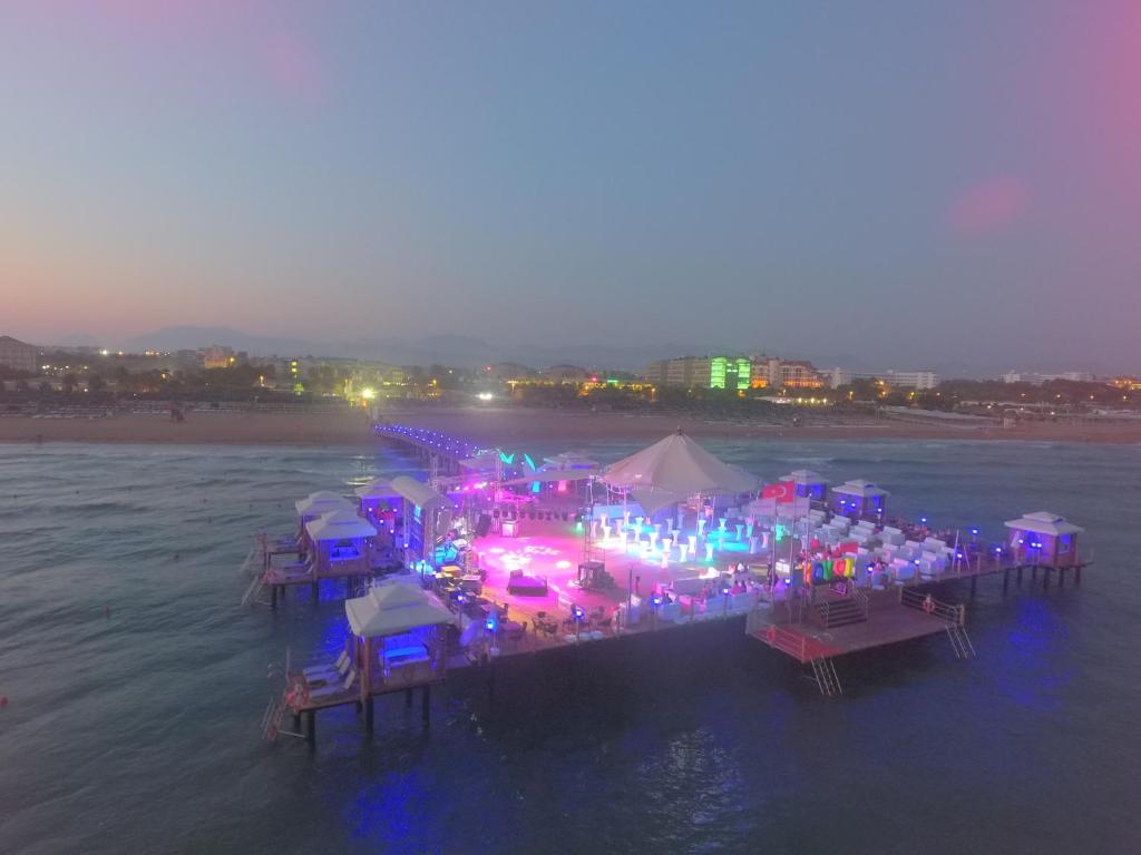 Krāsains klubs uz mola pie jūras, izgaismots ar dažādām gaismām, kas rada svinīgu atmosfēru. Fons attēlo pludmali un pilsētas apgaismojumu krēslā.