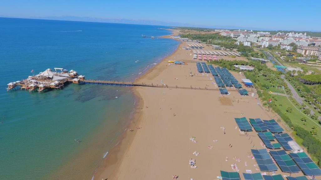 Pludmale ar zeltainu smiltīm, saulainu laiku un pludmales kafejnīcu, kas atrodas uz piestātnes. Fona ainavā redzami kūrorta viesnīcas un saules aizsargteltis.