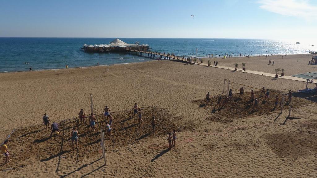 Pludmale ar cilvēkiem, kas spēlē pludmales volejbolu, un skats uz jūru ar laivu piestātni. Silta, saulaina diena, kur cilvēki izbauda ūdeni un smiltis.