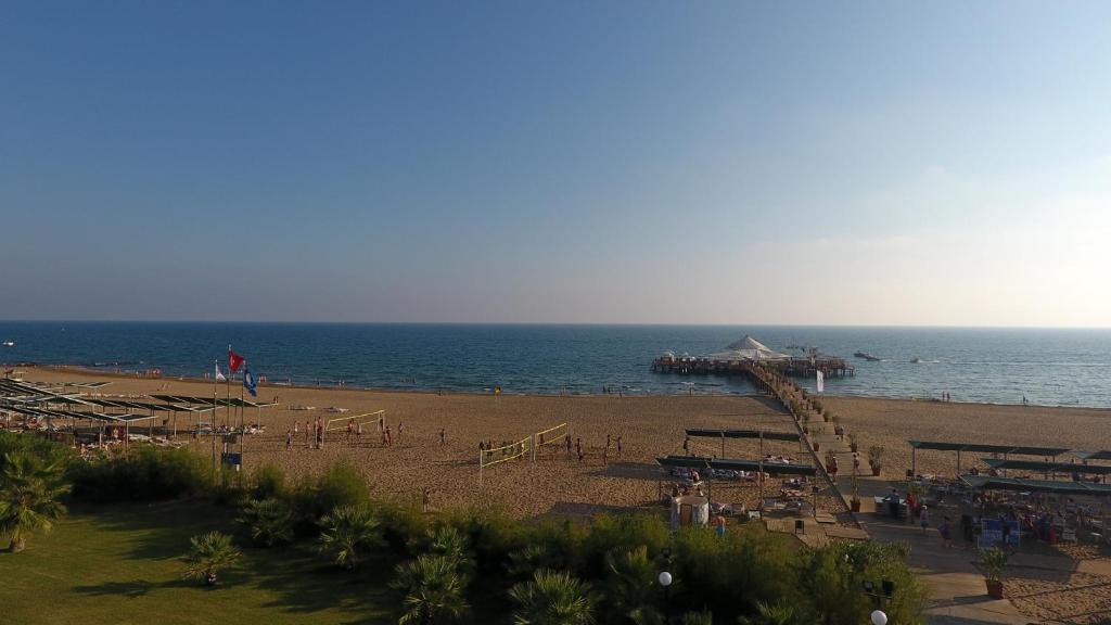 Pludmales ainava ar smilšainu krastu, sauļojošiem cilvēkiem un piestātni, kas ved uz jūru. Skats uz zilo ūdeni un skaidro debesi.