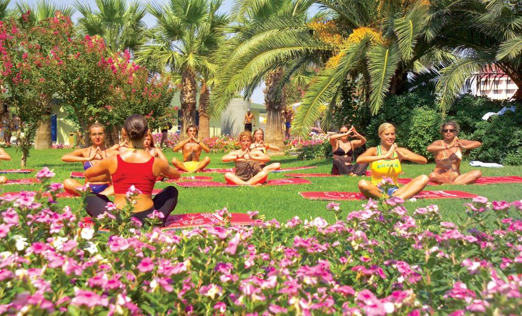 Jogas nodarbība dabā ar dalībniekiem, kas sēž uz paklājiem zālē, apstādīta ar ziediem un palmām. Visi praktizē elpošanas un meditācijas tehniku, baudot mierīgus dabu.