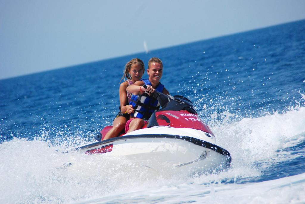 Divi cilvēki izbauda braucienu ar ūdens motociklu, priecājoties par saulainu laiku un viļņiem. Fons veido zila jūra un skaidrs debesis.