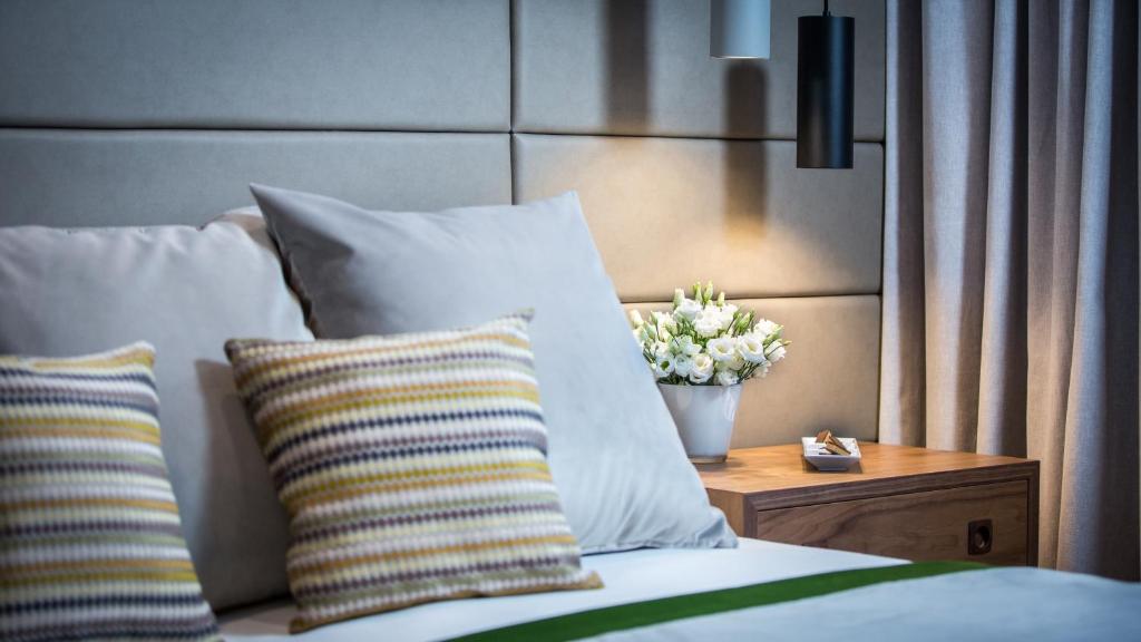 Mūsdienīgs guļamistabas interjers ar ērtām spilveniem un ziedu kompozīciju uz naktsgaldiņa. Mīkstas krāsas un apgaismojums rada relaksējošu atmosfēru.