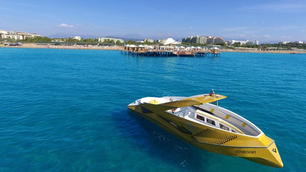 Dzeltenā laiva kuģo zilajos ūdeņos pie pludmales ar viesnīcām fona. Skats uz koka molu un pludmali.