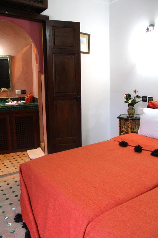 Mierīgā guļamistaba ar sarkanu segu, dekoratīvu galdiņu un piekļuvi vannas istabai. Interjerā ir koka durvis un ziedu kompozīcija uz galda.