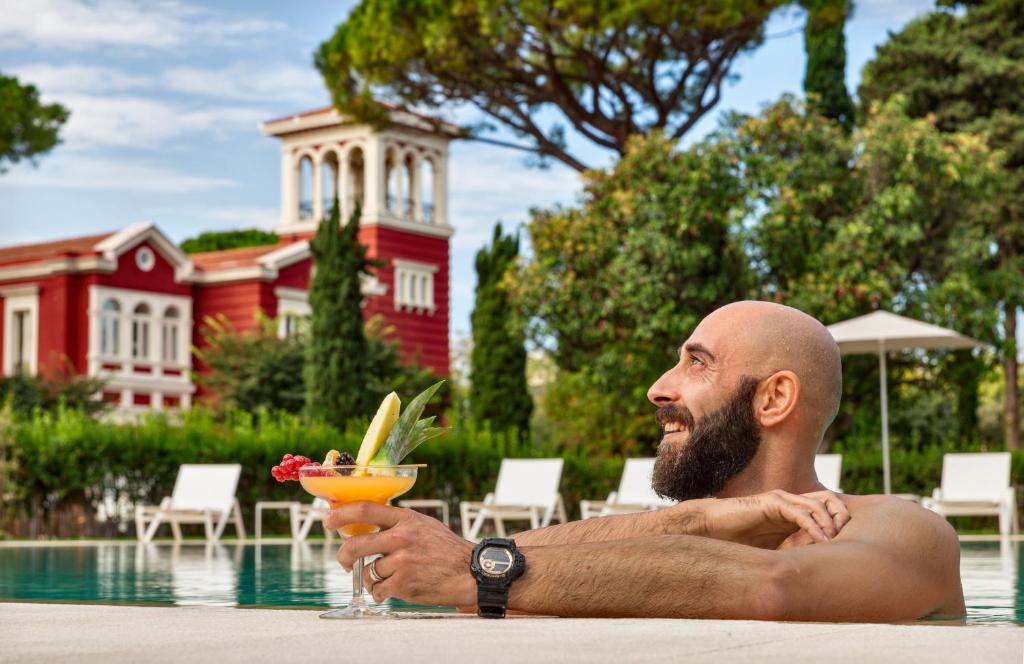 Vīrietis relaksējas pie baseina, turot rokās kokteili, kam ir augļi. Fons veido elegantā sarkanā ēka ar zaļiem kokiem un saulainu laiku.