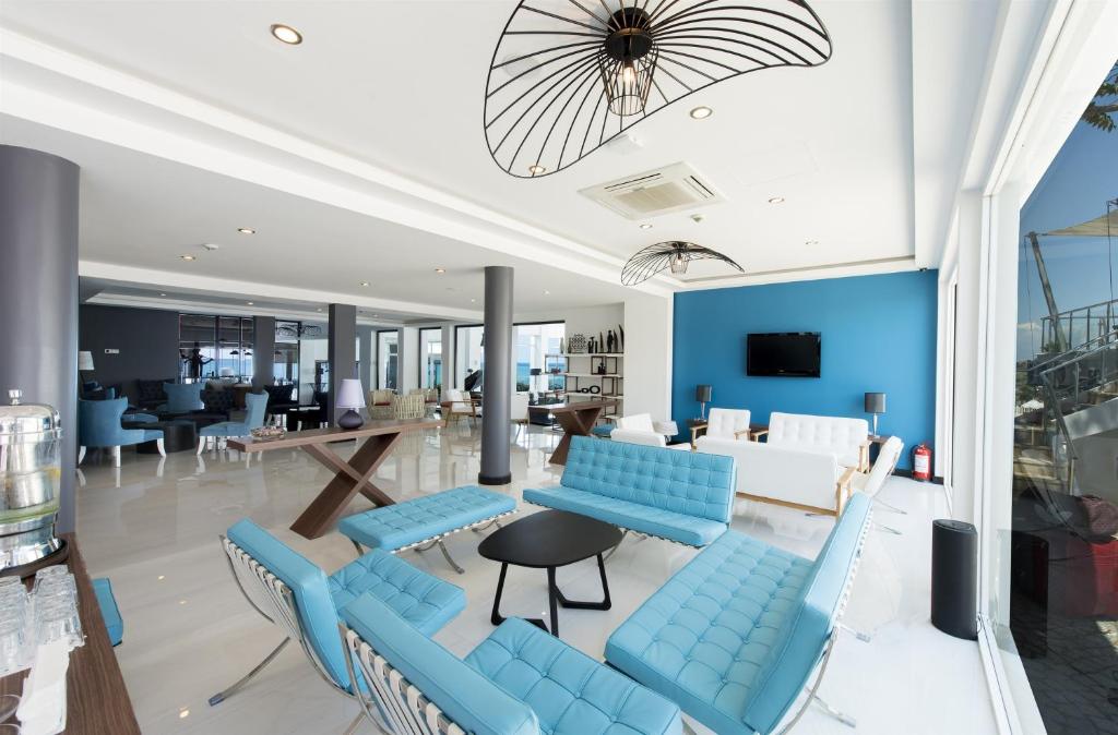 Modernā viesnīcas uzgaidāmā telpa ar gaišiem toņiem un stilīgu mēbelējumu, kas ietver zilo dīvānu un elegantu galdu. Telpa ir apgaismota ar pievilcīgām lampām un ir labi redzama caur lielajiem logiem.