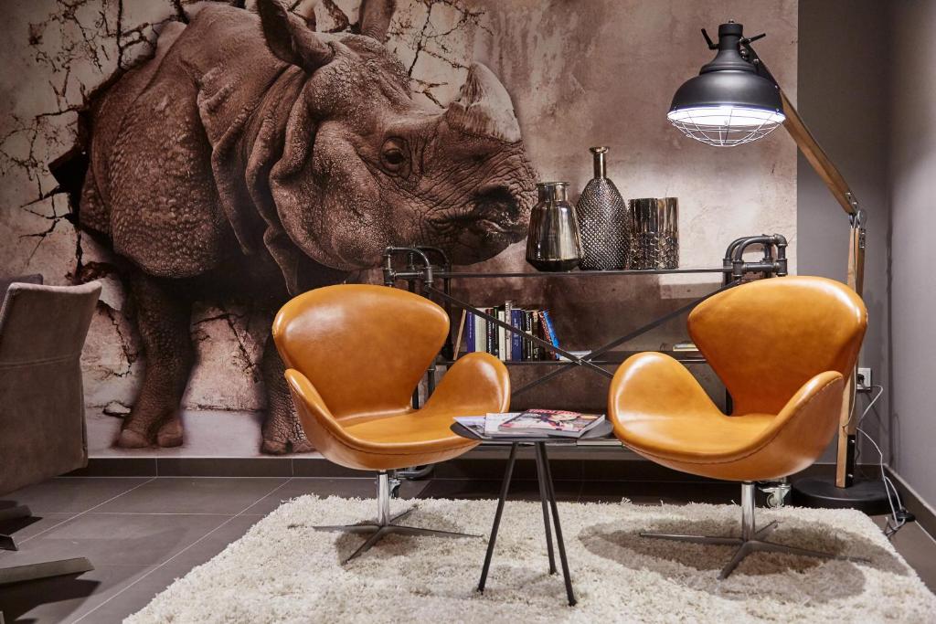 Mūsdienīga viesistaba ar divām oranžām krēslām, stikla galdu un aizraujošu rinoceros attēlu uz sienas. Interjerā ir eleganti aksesuāri un siltas krāsas, kas rada mājīgu atmosfēru.