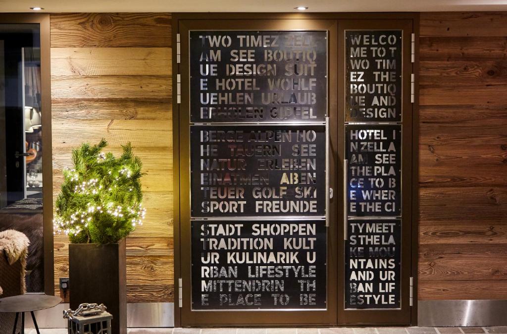 Mūsdienīgs viesnīcas interjers ar koka sienu un stikla durvīm, uz kurām redzami dažādi uzraksti. Dekorācija papildināta ar mazu gaismu iemirdzošu kokaugu.