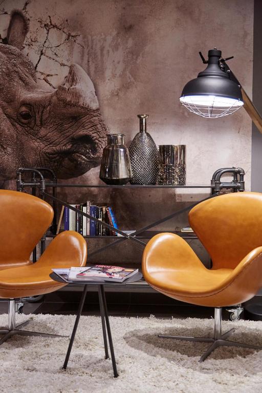 Stilīgs interjers ar divām oranžām krēslām, melnu galdiņu un dekoratīvām vāzēm uz cauruļu plaukta, aiz kura redzama rinocera attēls uz sienas.