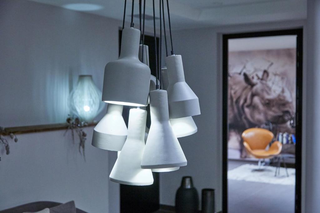 Mūsdienīga gaismekļu grupa, kas izgatavota no pelēka materiāla, rada siltu un mājīgu atmosfēru interjerā. Fona attēlā redzama dīvāna zona ar krēslu un mākslas darbu uz sienas.