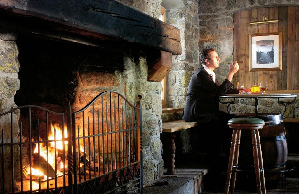 Eleganta vīrieša atpūta pie kamīna, baudot dzērienu un cigāru mājīgā akmens interjerā. Iedegta uguns rada siltu un relaksējošu atmosfēru.