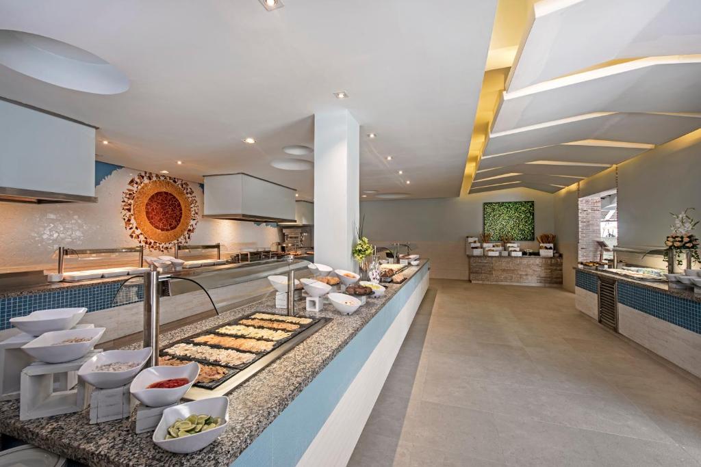 Mūsdienīgs restorāns ar plašu bufeti, noformēts ar gaišām krāsām un elegantām detaļām. Redzami dažādi ēdieni un dekoratīvās sastāvdaļas, kas veido pievilcīgu atmosfēru.