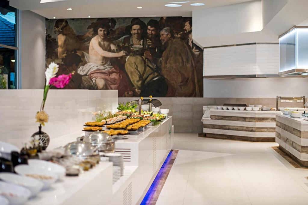 Mūsdienīga bufete ar dažādiem ēdieniem, ko rotā ziedi, un aizkadra glezna, kas piešķir telpai māksliniecisku atmosfēru.